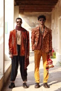 Two men in bold Indo-western outfits with patterned jackets, scarves and trousers, blending ethnic wear with modern streetwear style